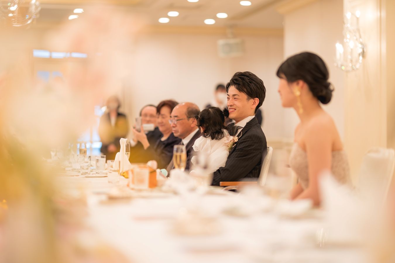 ANAクラウンプラザホテル松山でご結婚式を挙げられたお客様の写真 ご家族様の乾杯のご挨拶を聞いている笑顔のタキシード姿の新郎様とカラードレス姿の新婦様の横顔